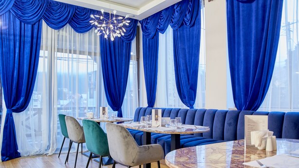 Un espace de restauration moderne avec une table ronde en marbre, des sièges rembourrés en tissu bleu et une vue sur la ville grâce à de grandes fenêtres.