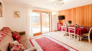 Een woonkamer met een bank, een eettafel met stoelen, een aan de muur bevestigde televisie en een schuifdeur naar een buitenruimte.