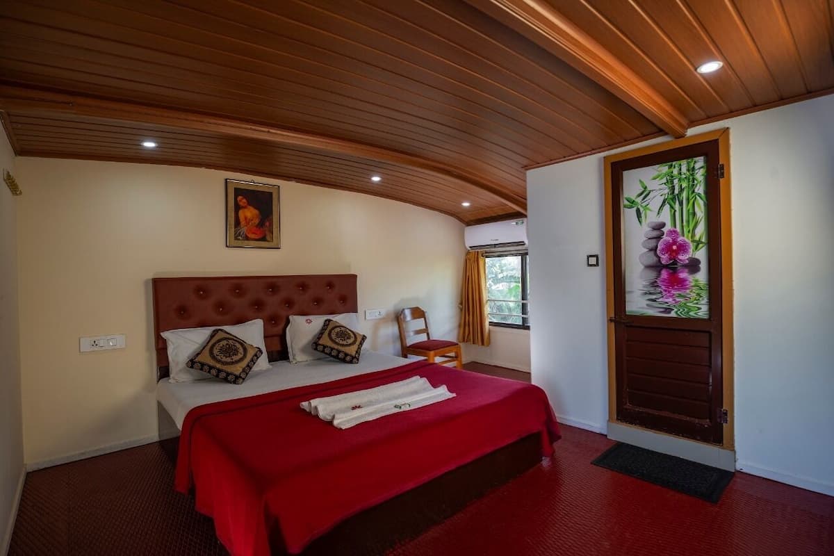 A hotel room with a wooden ceiling, a bed with a red bedspread, a painting on the wall, and a door with a floral design.