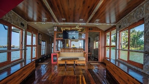 Interior de barco con pisos de madera, zona de estar central y amplios ventanales con vistas al agua.