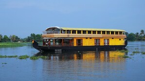 Una casa flotante amarilla y negra con cubierta superior protegida, flotando sobre aguas tranquilas y con vegetación al fondo.