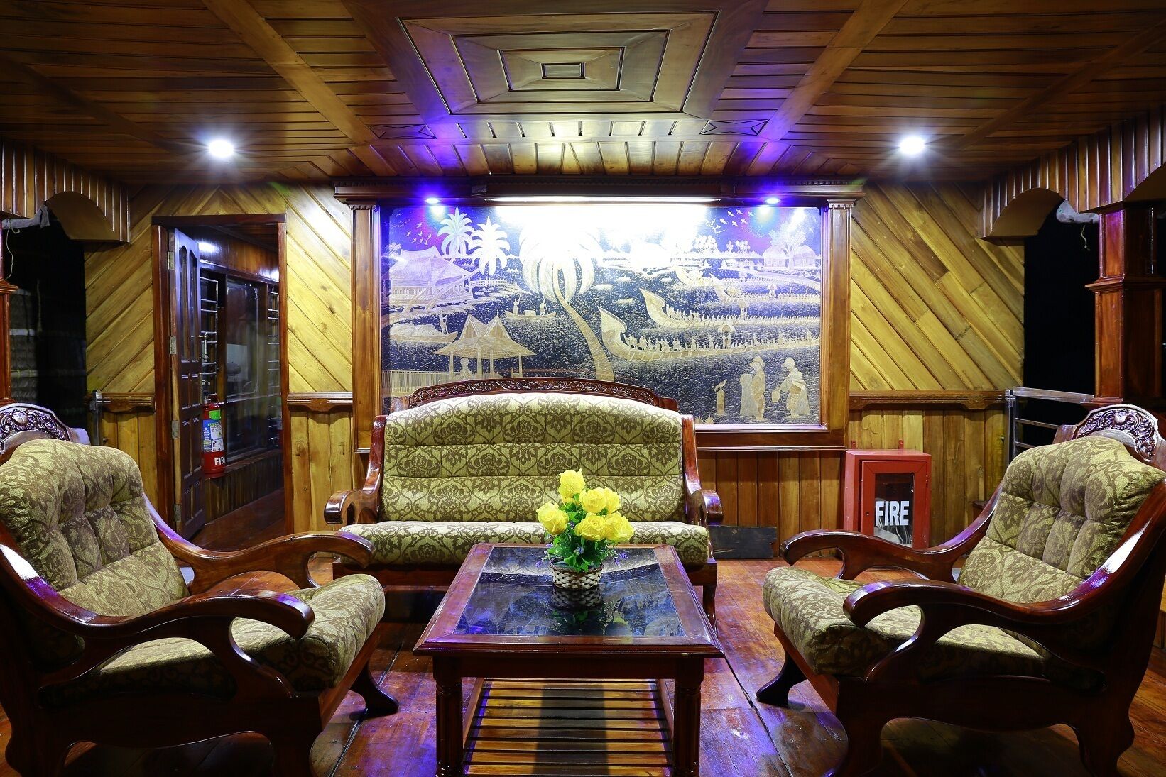 Una habitació amb un sofà estampat, dues butaques, una taula de cafè de vidre amb un ram de flors grogues i un mural gran que representa una escena tradicional.