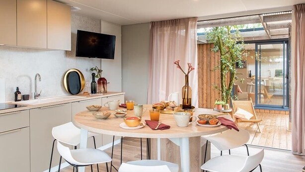 Una cocina moderna con comedor, que incluye una mesa redonda con sillas, un televisor montado sobre la encimera y un amplio ventanal con cortinas.