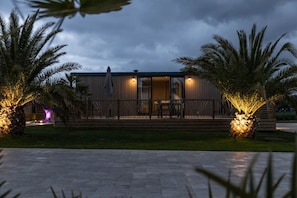 A modern house with a wooden deck, surrounded by palm trees and a paved walkway. - M&V Resort (Langrune-sur-Mer)
