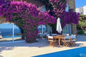 Uma área à beira da piscina com mesa e cadeiras, rodeada por exuberante bougainvillea roxa.