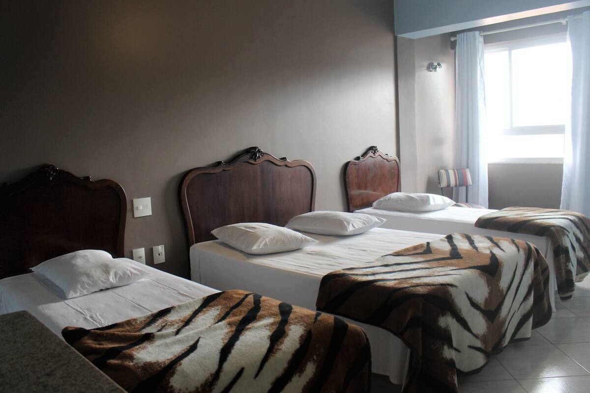 A hotel room with two beds, each with a wooden headboard and white bedding. There is a window with curtains, a bedside table with a lamp, and a wall-mounted light switch.
