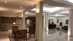 Een ruime lobby met een receptiebalie, houten stoelen, een bank en een grote pilaar.