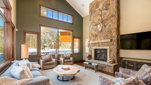 A spacious living room with a stone fireplace, comfortable seating, and large windows offering a view of a mountain cabin. - Short Walk to River Run, Private Hot Tub, Tons of Sunshine and Natural Light (Keystone)