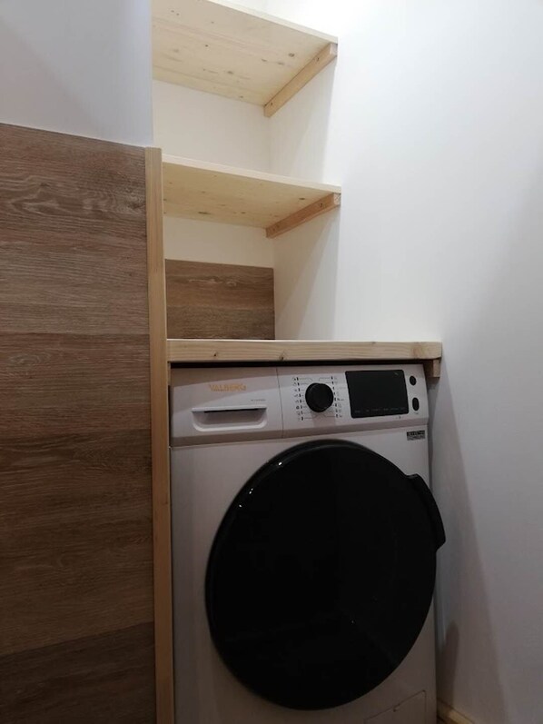 Un lavadero pequeño con lavadora de carga frontal, repisas de madera arriba y un gabinete de madera empotrado.