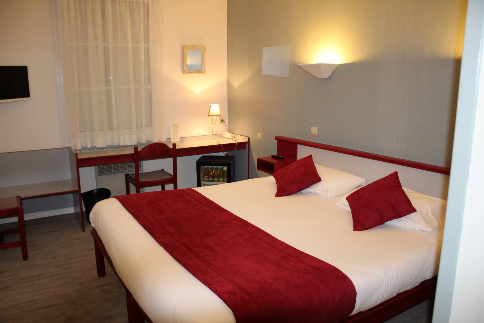 A hotel room with a bed, a desk, a chair, a small table, a mirror, and a wall-mounted light.
