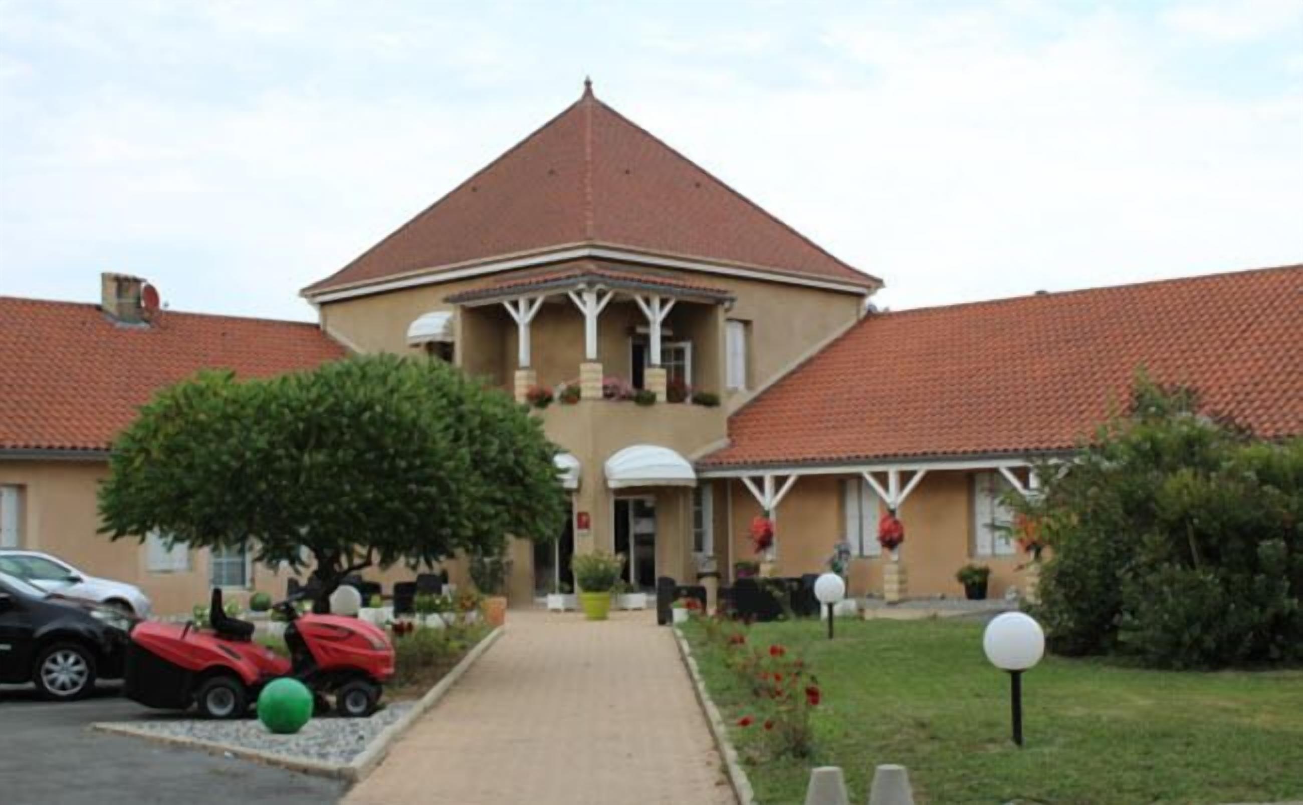 A two-story building with a red-tiled roof and a central balcony. There are cars parked in front and a pathway leading to the entrance.