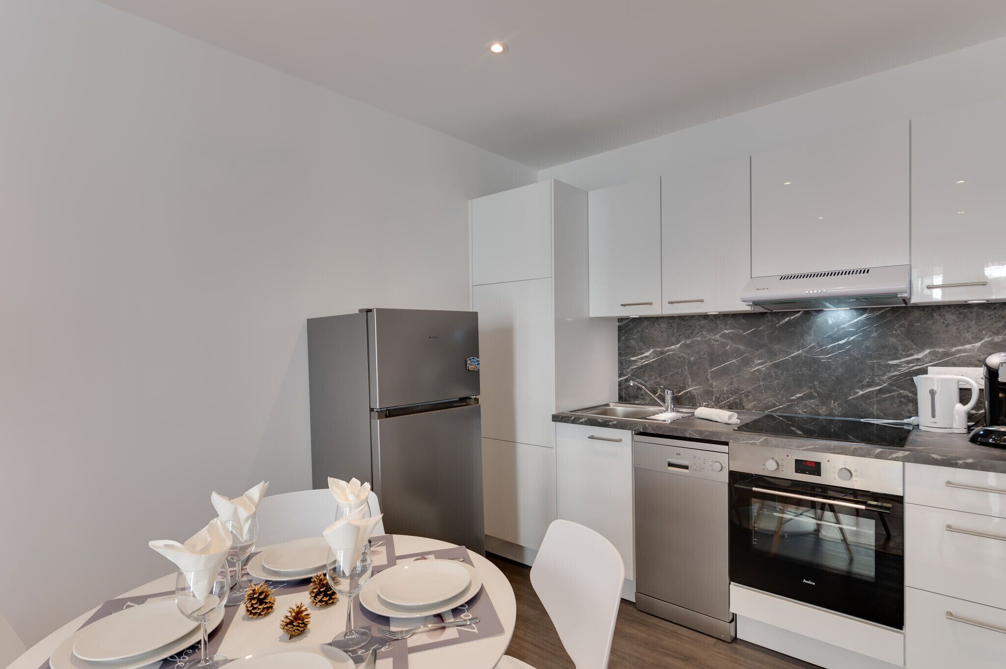 A modern kitchen with white cabinets, a stainless steel refrigerator, a built-in oven, and a marble countertop.