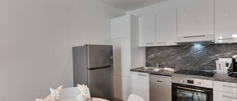 A modern kitchen with white cabinets, a stainless steel refrigerator, a built-in oven, and a marble countertop.