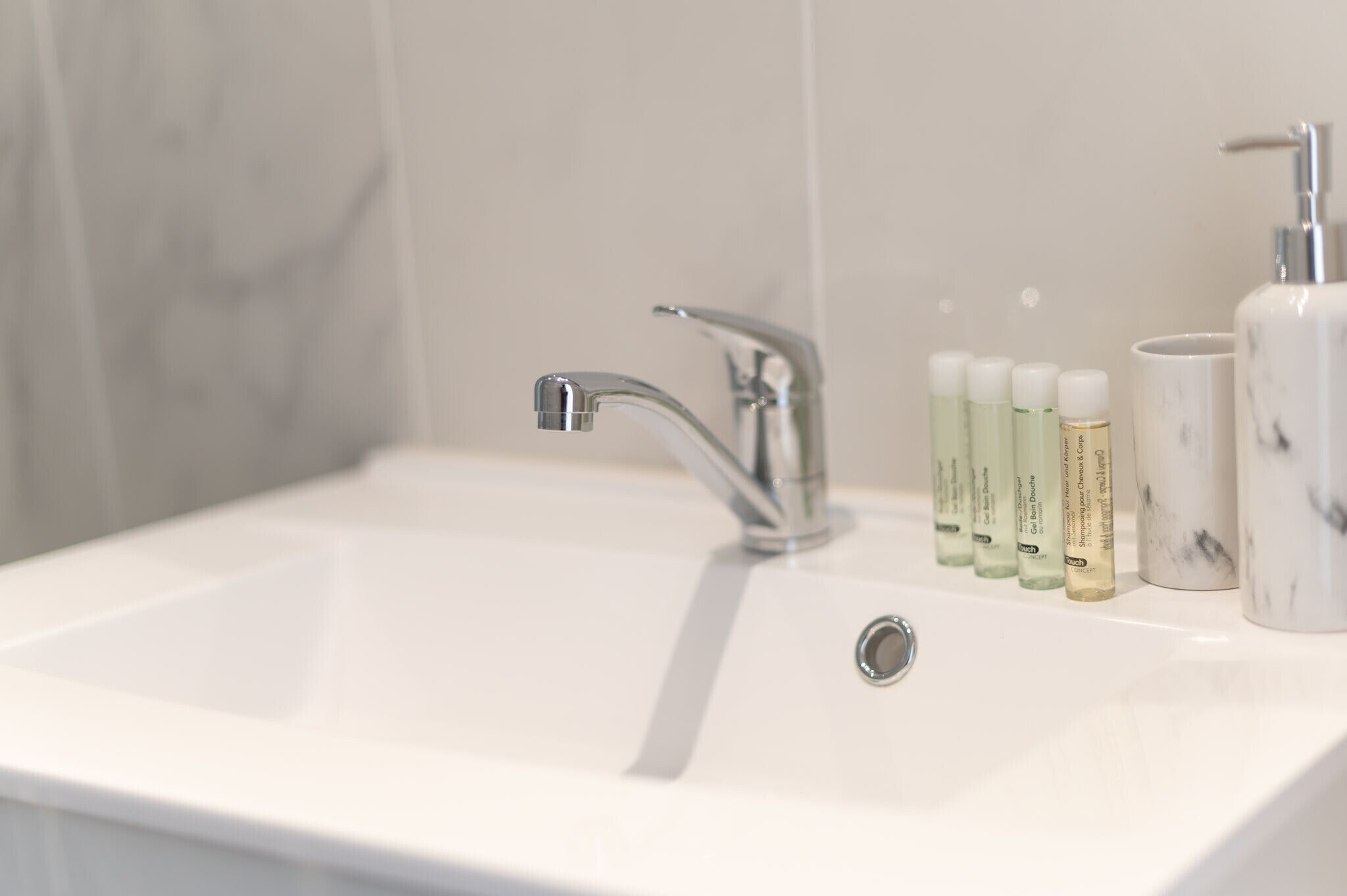 A bathroom sink with a modern faucet, a marble countertop, and a row of toiletries including a soap dispenser and lotion tubes.