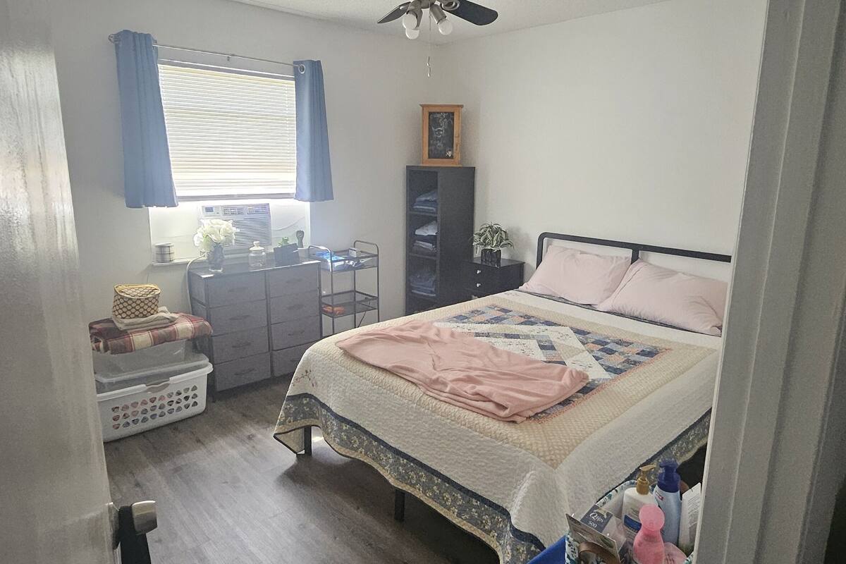 A bedroom with a bed, a window with curtains, a dresser, a shelf with items, and a ceiling fan.