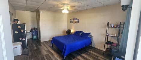 A bedroom with a bed, a ceiling fan, a wall-mounted shelf, and a small kitchenette area.