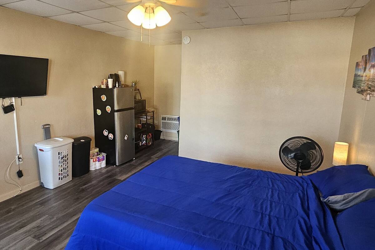 A room with a bed, a desk with a chair, a television, a refrigerator, a fan, and a wall clock.