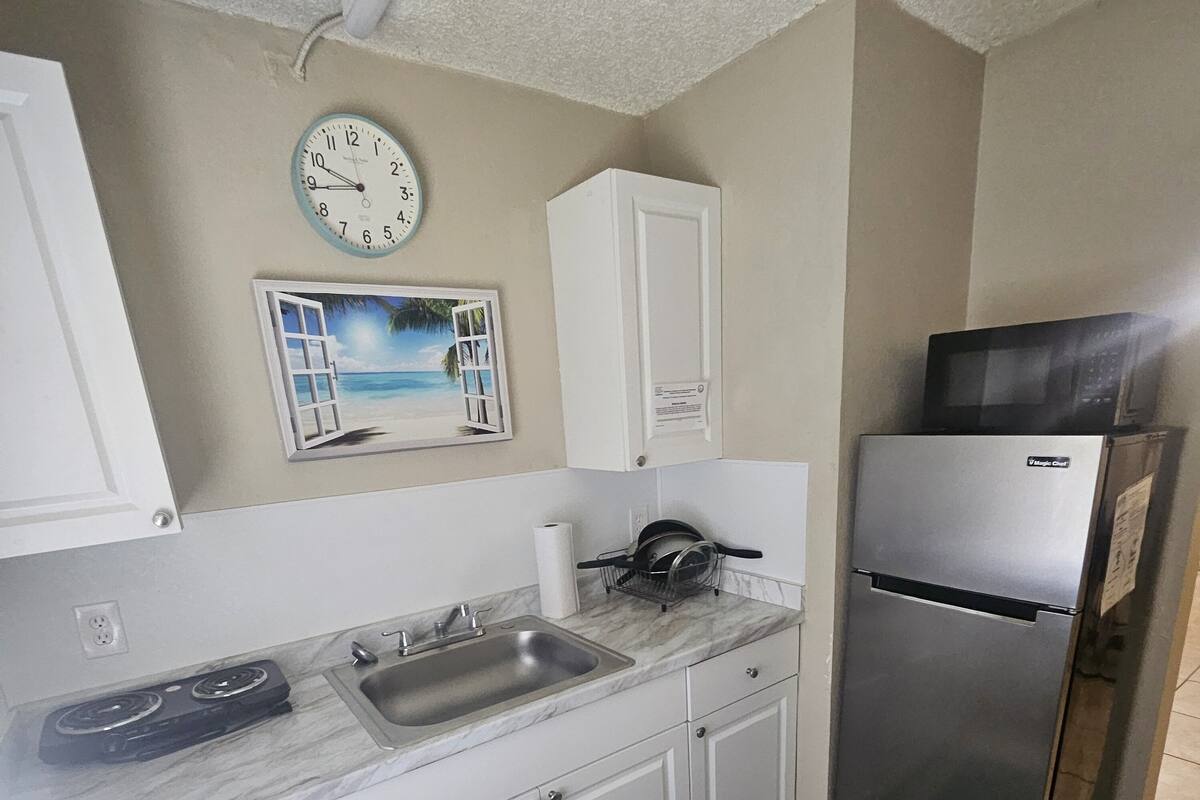 A kitchen with white cabinets, a stainless steel refrigerator, a microwave, a sink, and a clock on the wall.