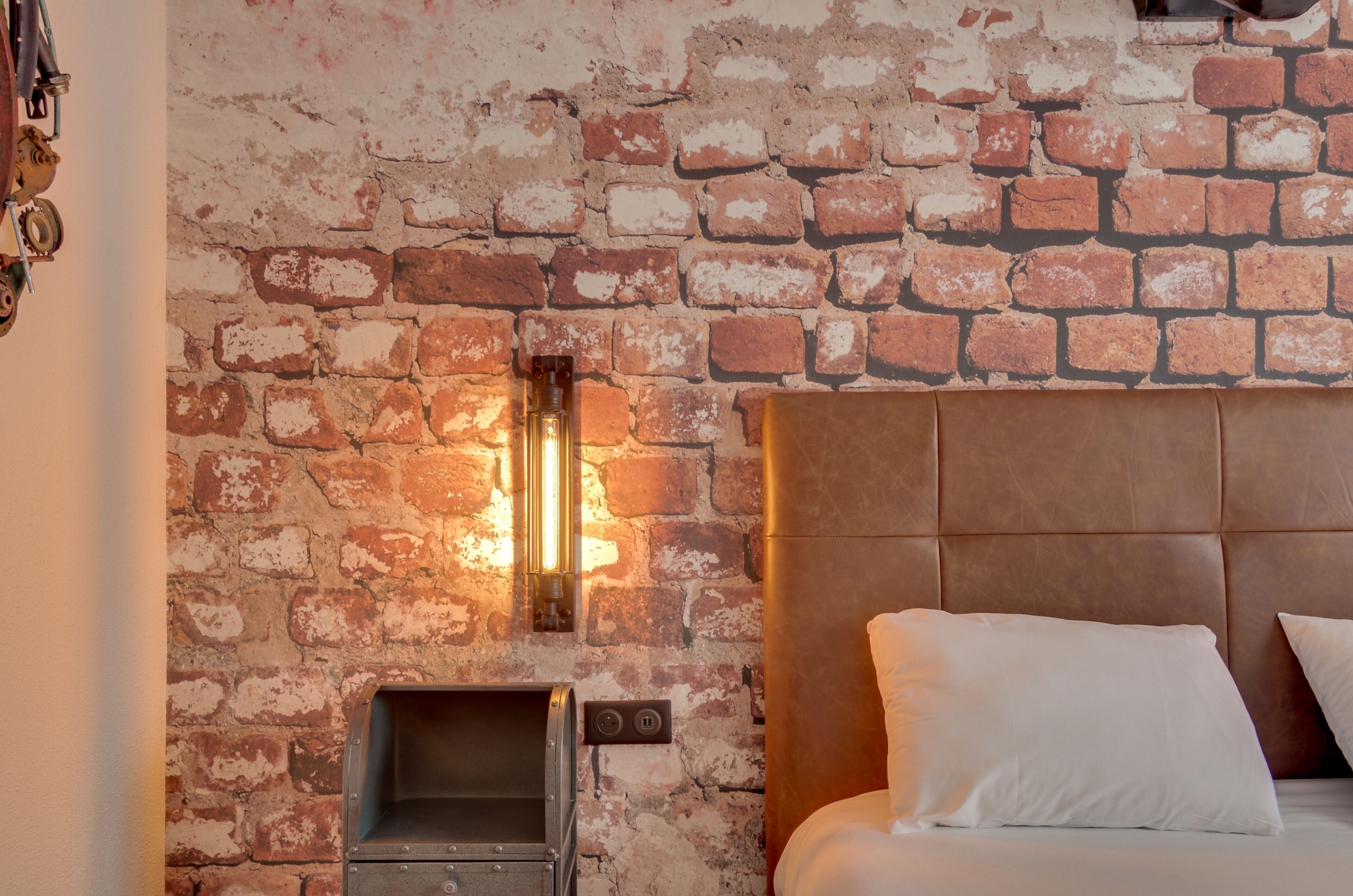 A room with a brick wall, a bed with a headboard, and a wall-mounted light fixture.