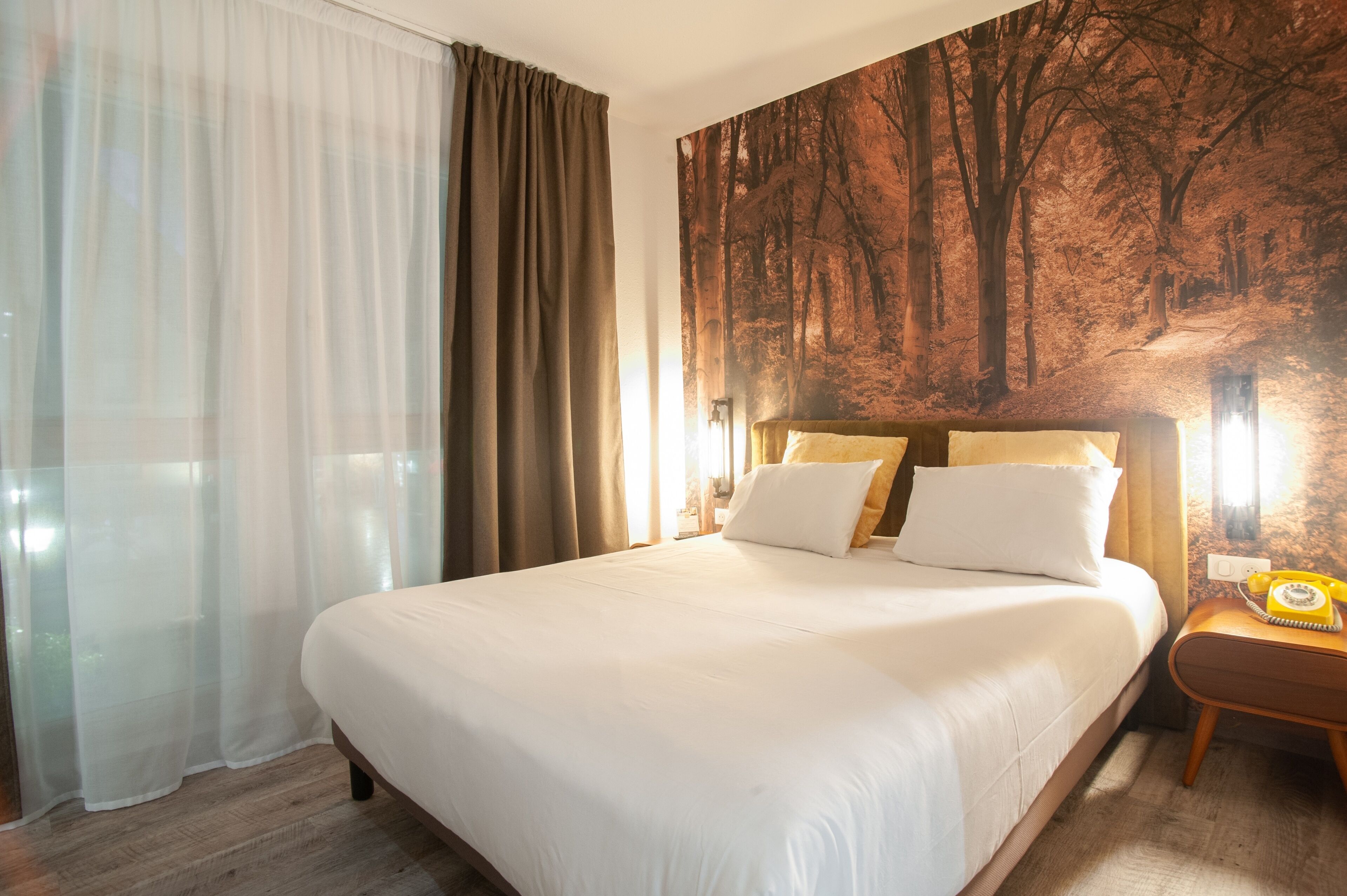 A hotel room with a large bed, a nightstand with a telephone, and a forest-themed wall mural.