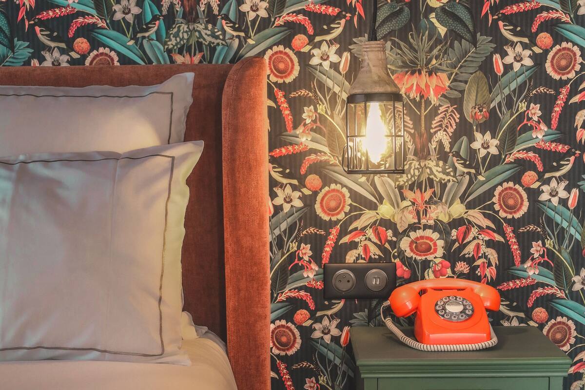 Ein Schlafzimmer mit blumigem Wandpapier, einem Bett mit weißer Bettwäsche, einem grünen Nachttisch mit Wählscheibe und einer Pendelleuchte.