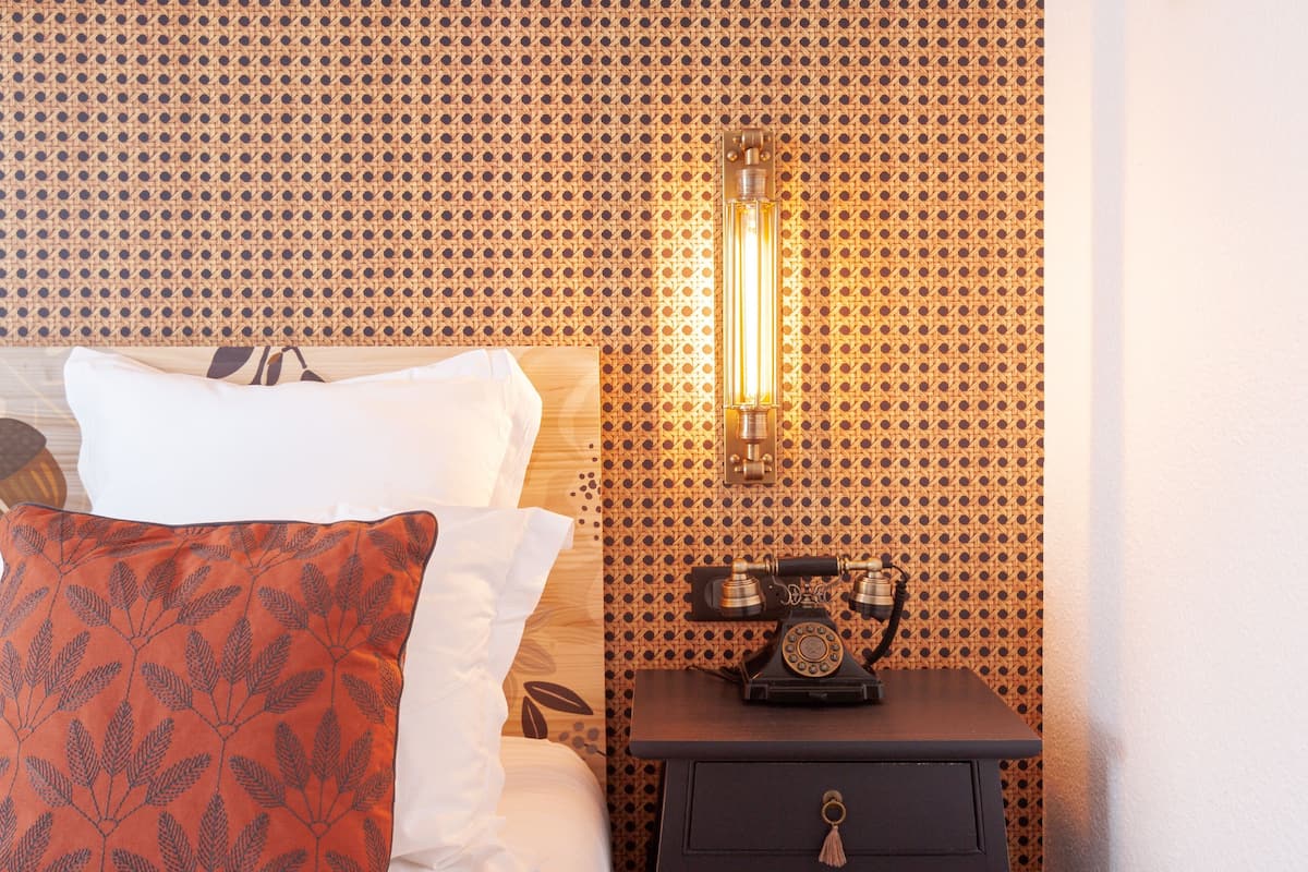 Ein ordentlich bezogenes Bett mit weißen und roten Kissen, ein altes Telefon auf einem Nachttisch und eine Wand mit geometrischem Muster.