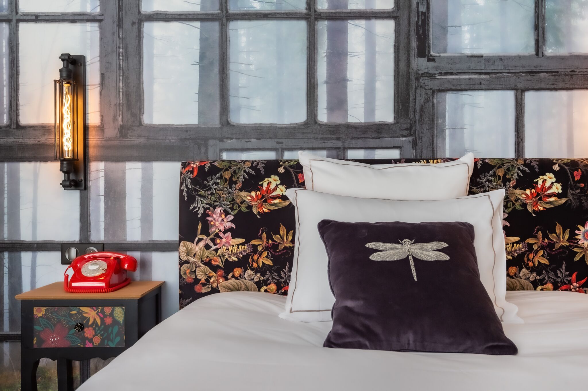 Un dormitorio con un cabecero estampado con motivos florales, un teléfono giratorio rojo sobre una mesita de noche y una ventana con vistas a árboles.