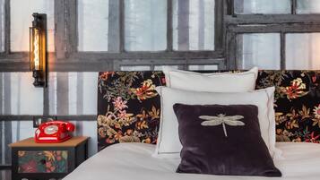 Un dormitorio con un cabecero estampado con motivos florales, un teléfono giratorio rojo sobre una mesita de noche y una ventana con vistas a árboles.