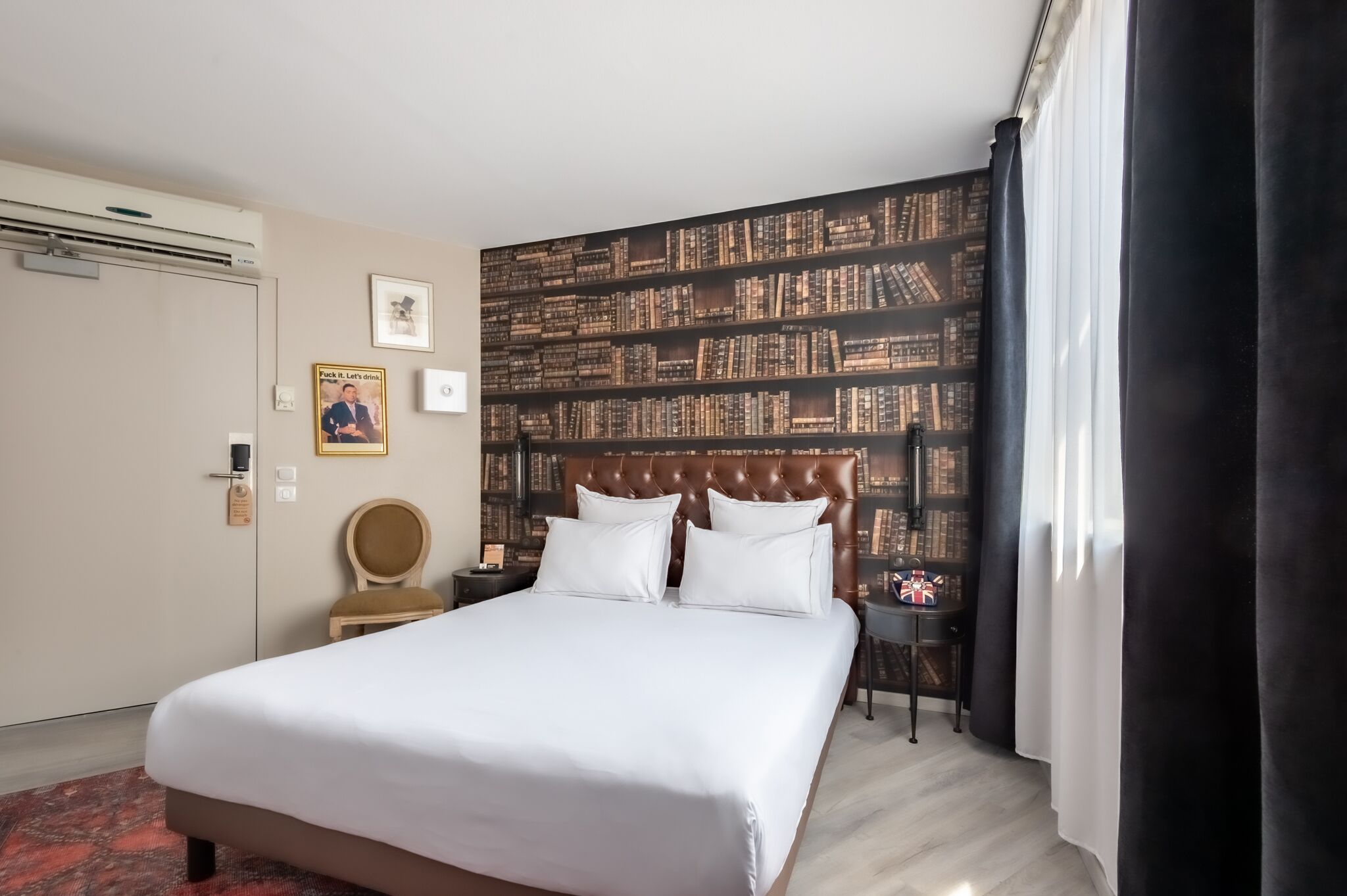 A hotel room with a large bed, a bookshelf wall, a wooden chair, a nightstand, and a window with curtains.