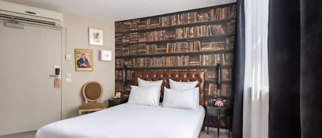 A hotel room with a large bed, a bookshelf wall, a wooden chair, a nightstand, and a window with curtains.