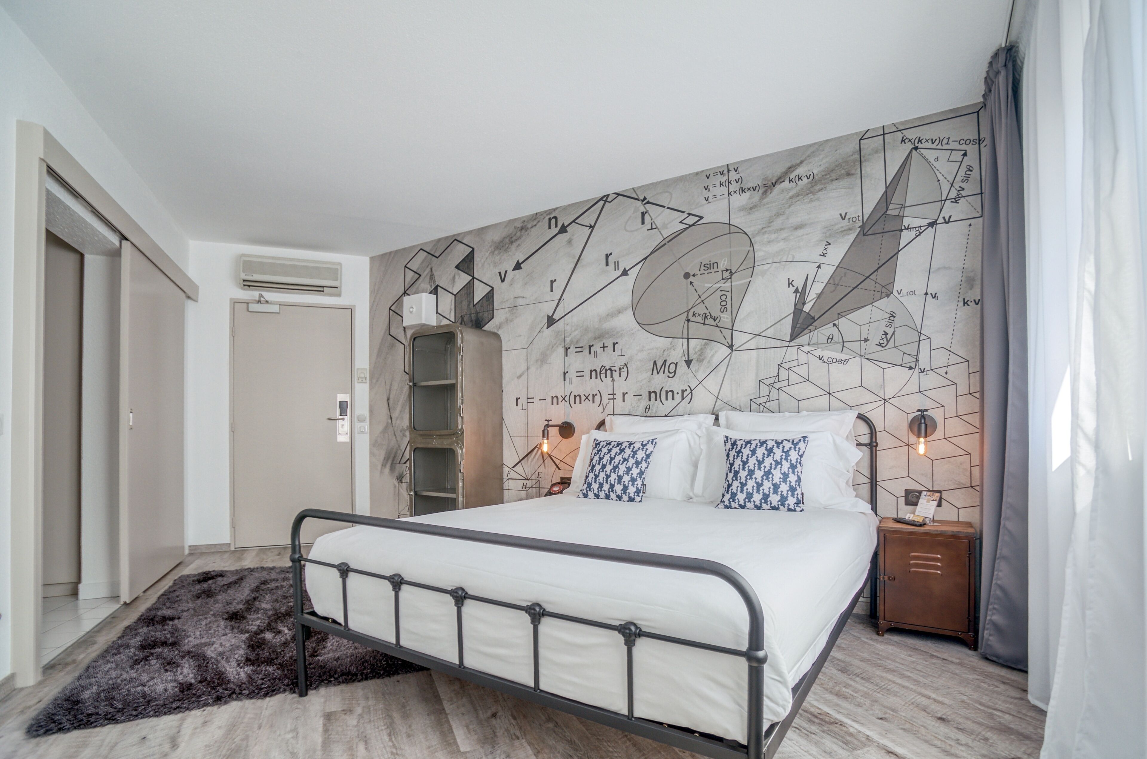 A bedroom with a bed, a nightstand, a wall-mounted air conditioner, a door, and a patterned rug.