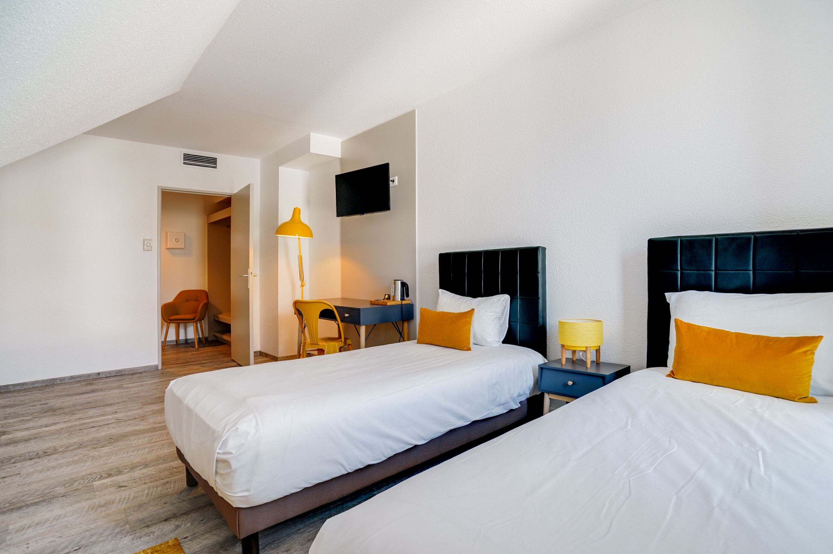 A hotel room with two beds, a desk, a chair, a TV, and a lamp.