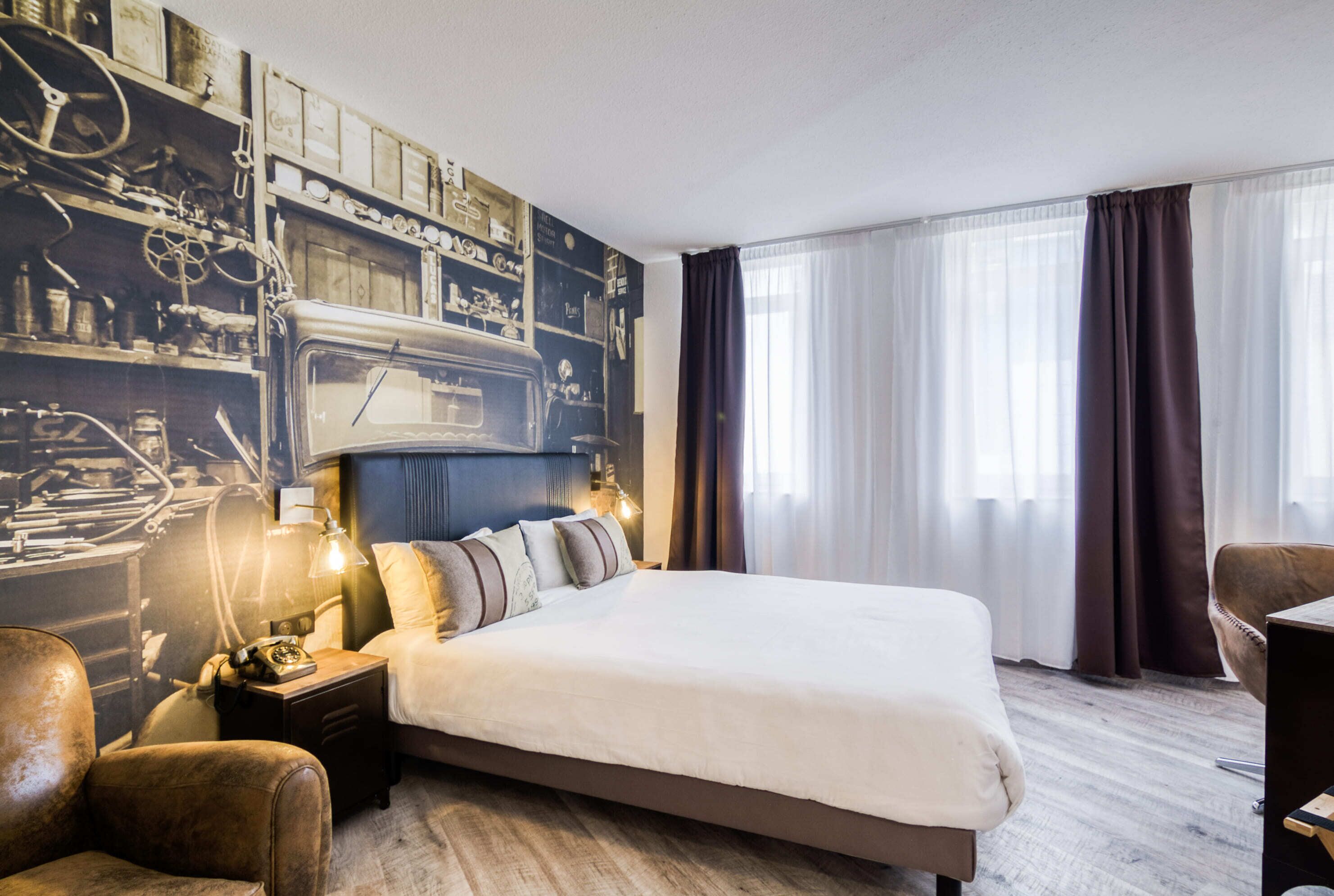 Hotellihuone, jossa on suuri sänky, yöpöytä lampun kanssa, vanhanaikainen puhelin ja seinä, jossa on mekaaninen kuvio.