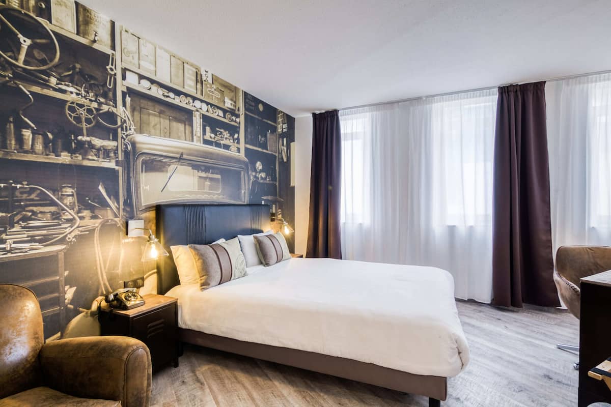 Hotellihuone, jossa on suuri sänky, yöpöytä lampun kanssa, vanhanaikainen puhelin ja seinä, jossa on mekaaninen kuvio.