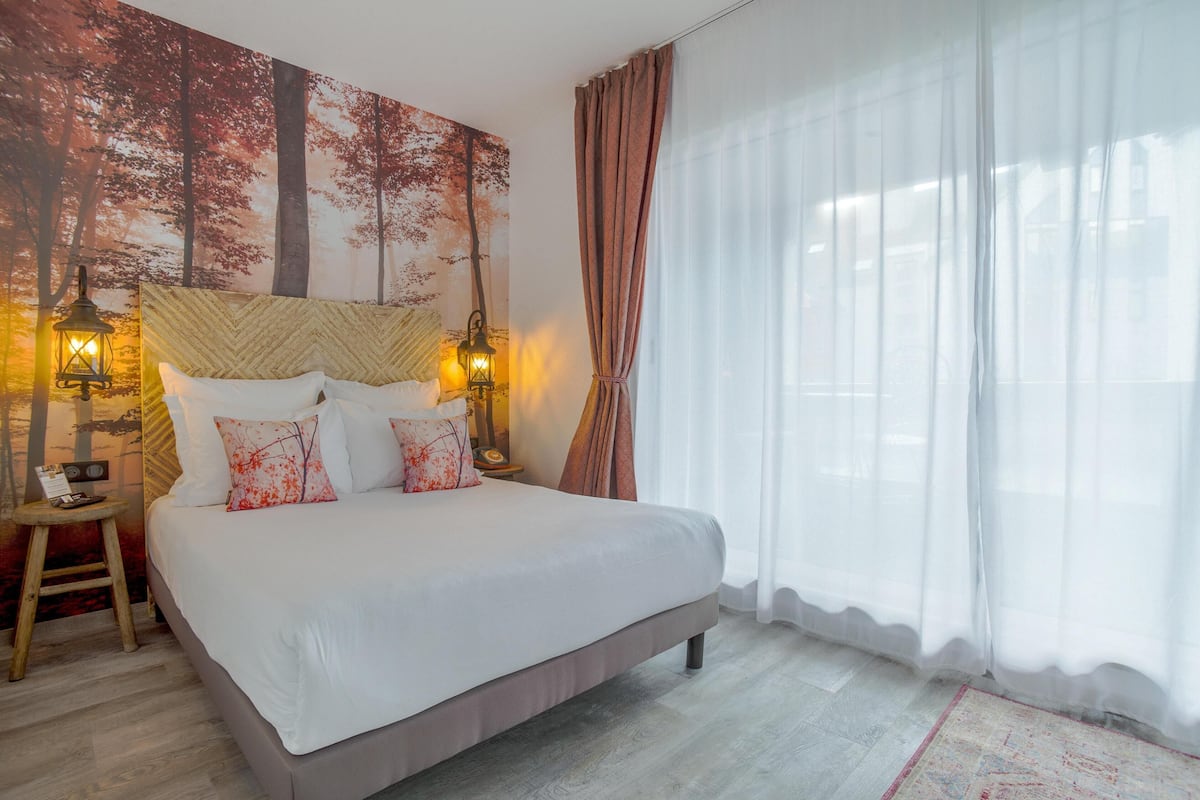 Ein ordentlich bezogenes Bett mit weißen Leinen und dekorativen Kissen, ein Nachttisch aus Holz mit einer Lampe und ein großes Fenster mit transparenten Vorhängen.