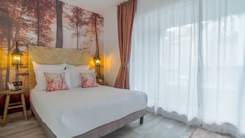 Ein ordentlich bezogenes Bett mit weißen Leinen und dekorativen Kissen, ein Nachttisch aus Holz mit einer Lampe und ein großes Fenster mit transparenten Vorhängen.