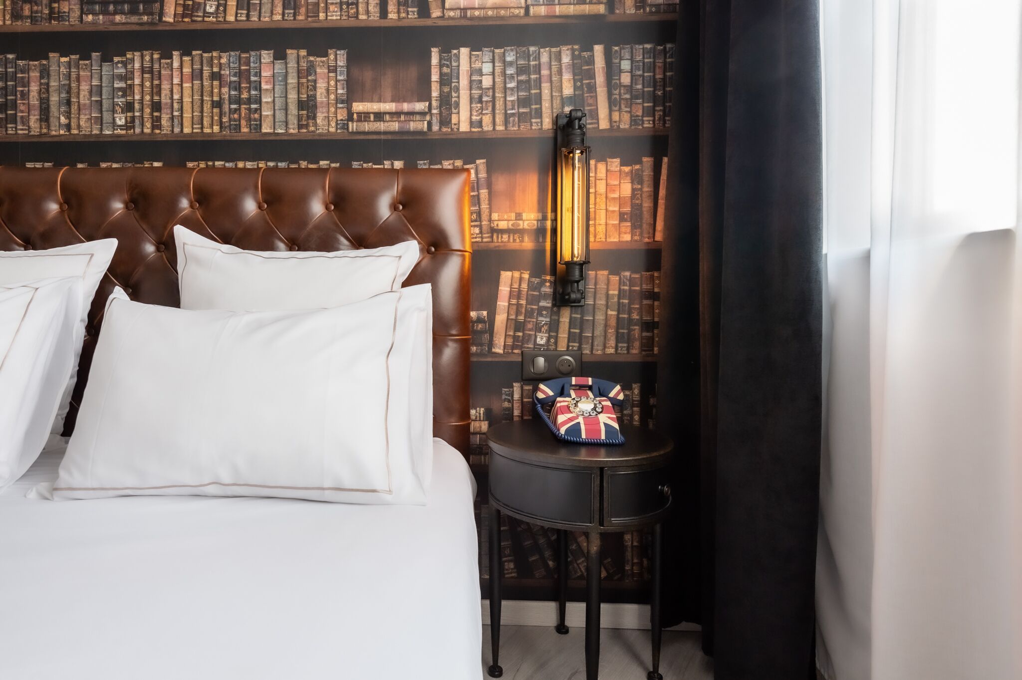 A bedroom with a large bed, a bookshelf, a small table with a lamp, and a window with curtains.