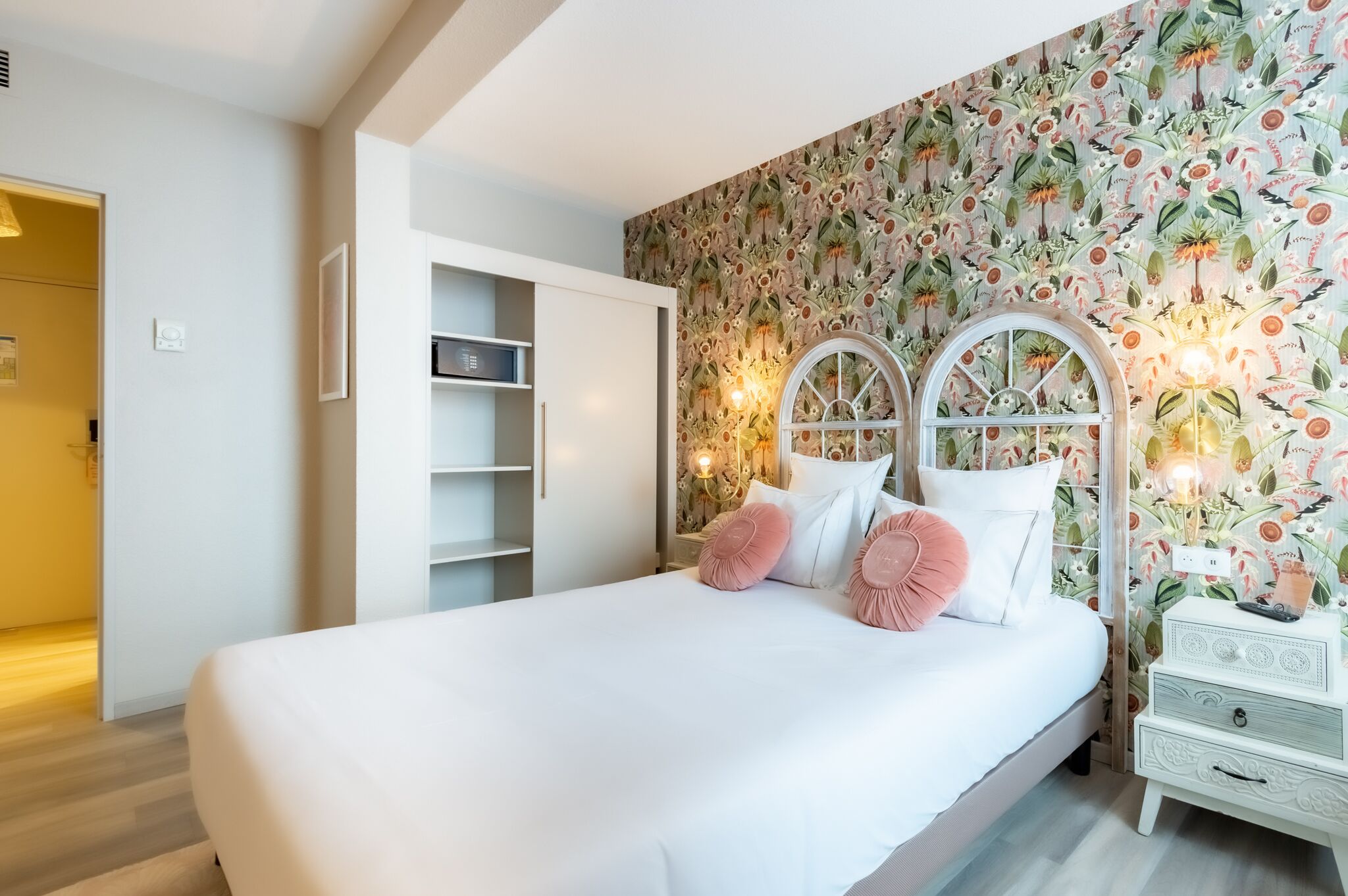 Een slaapkamer met een groot bed, witte muren, bloemenbehang en een witte nachtkastje met een lamp.