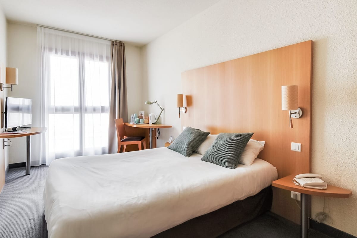 Ein Hotelzimmer mit einem großen Bett, einem Schreibtisch mit Stuhl, einer Lampe und einem Telefon auf einem Nachttisch.