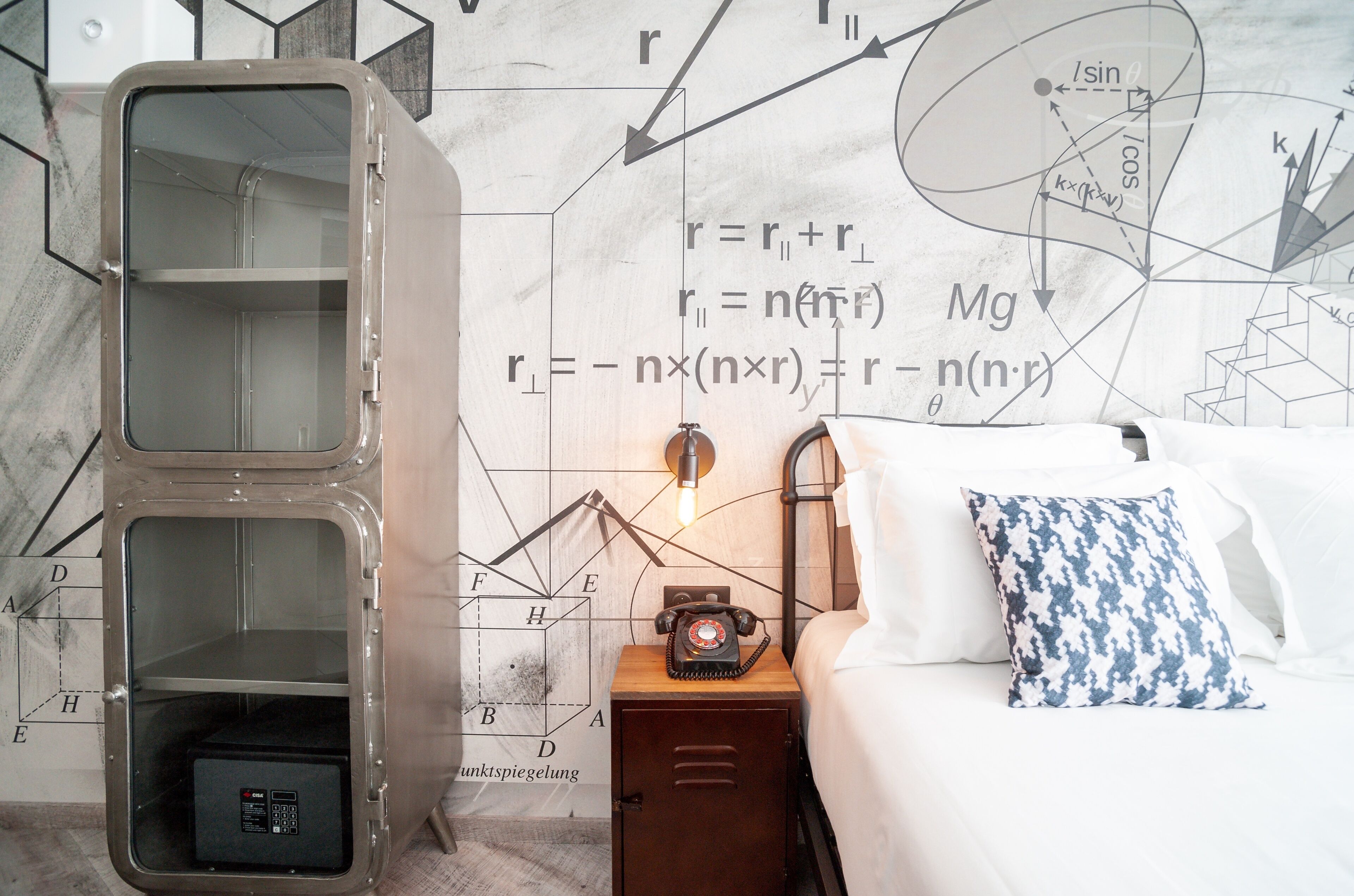 Una habitación con una caja fuerte grande, una cama, una mesita de noche con lámpara, y una pared con ecuaciones y diagramas matemáticos.