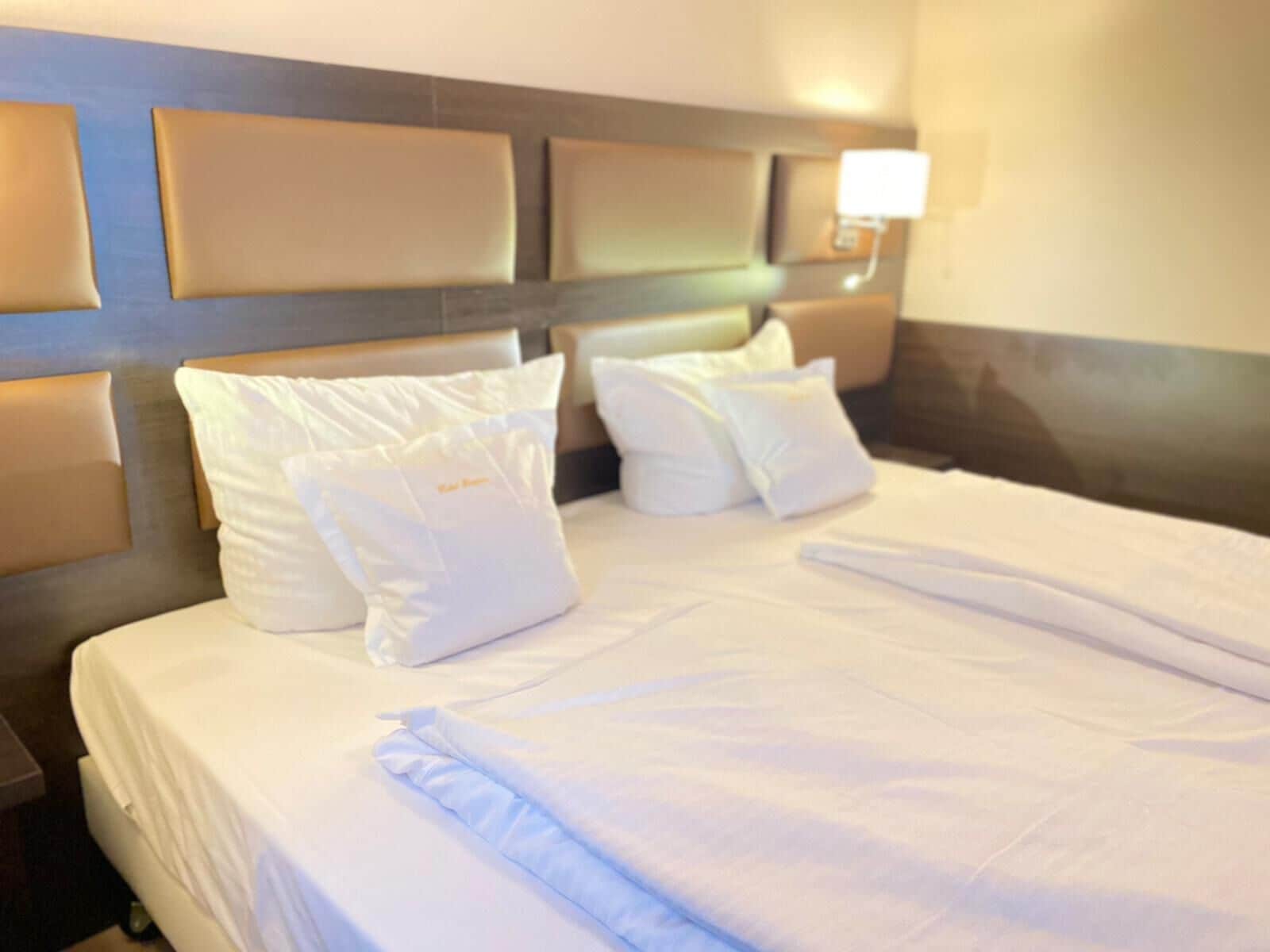 Una cama bien tendida con sábanas y almohadas blancas, un cabecero con una lámpara, y una luz montada en la pared.