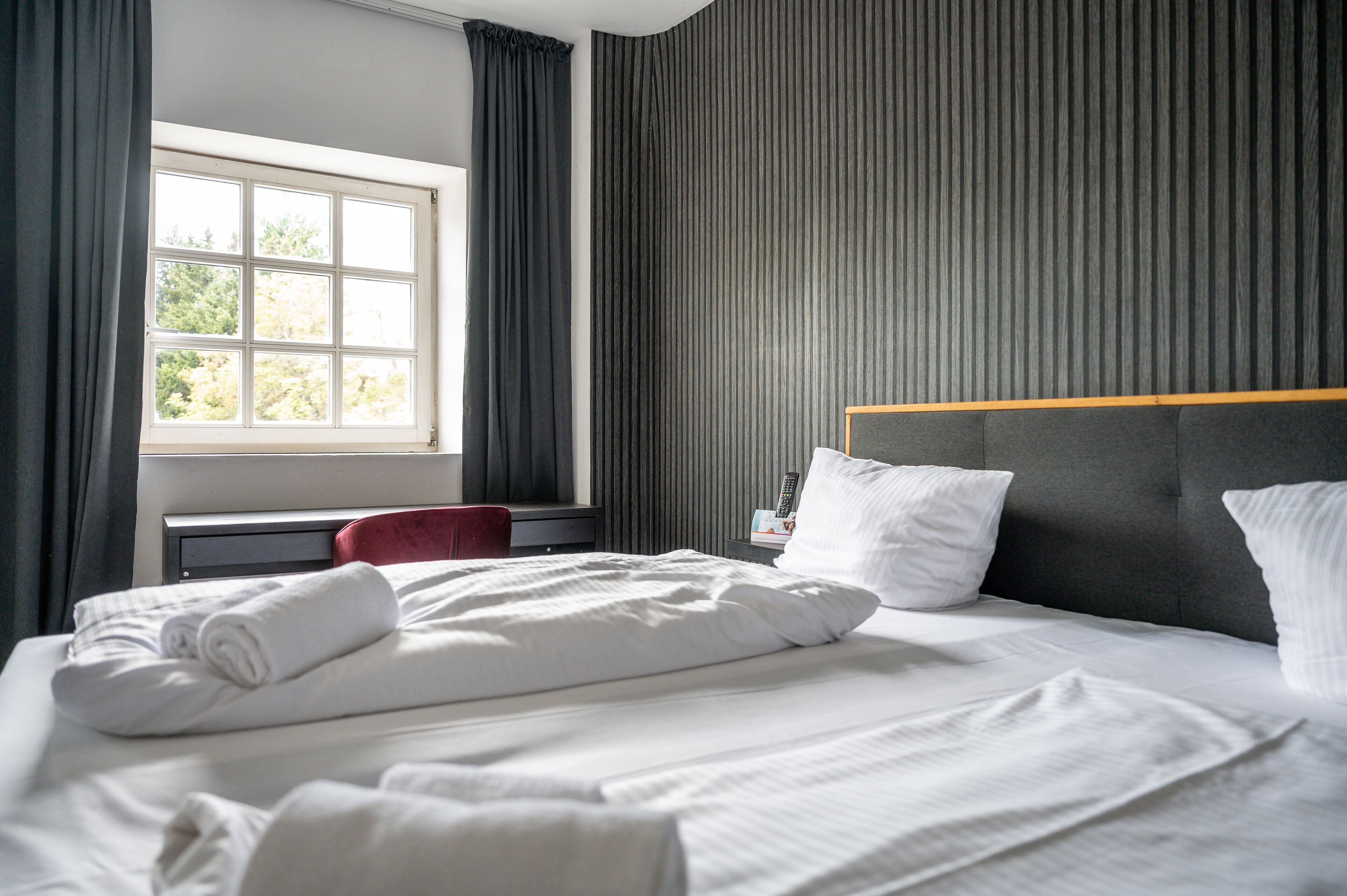A neatly made bed with white linens, a wooden headboard, and a window with curtains.