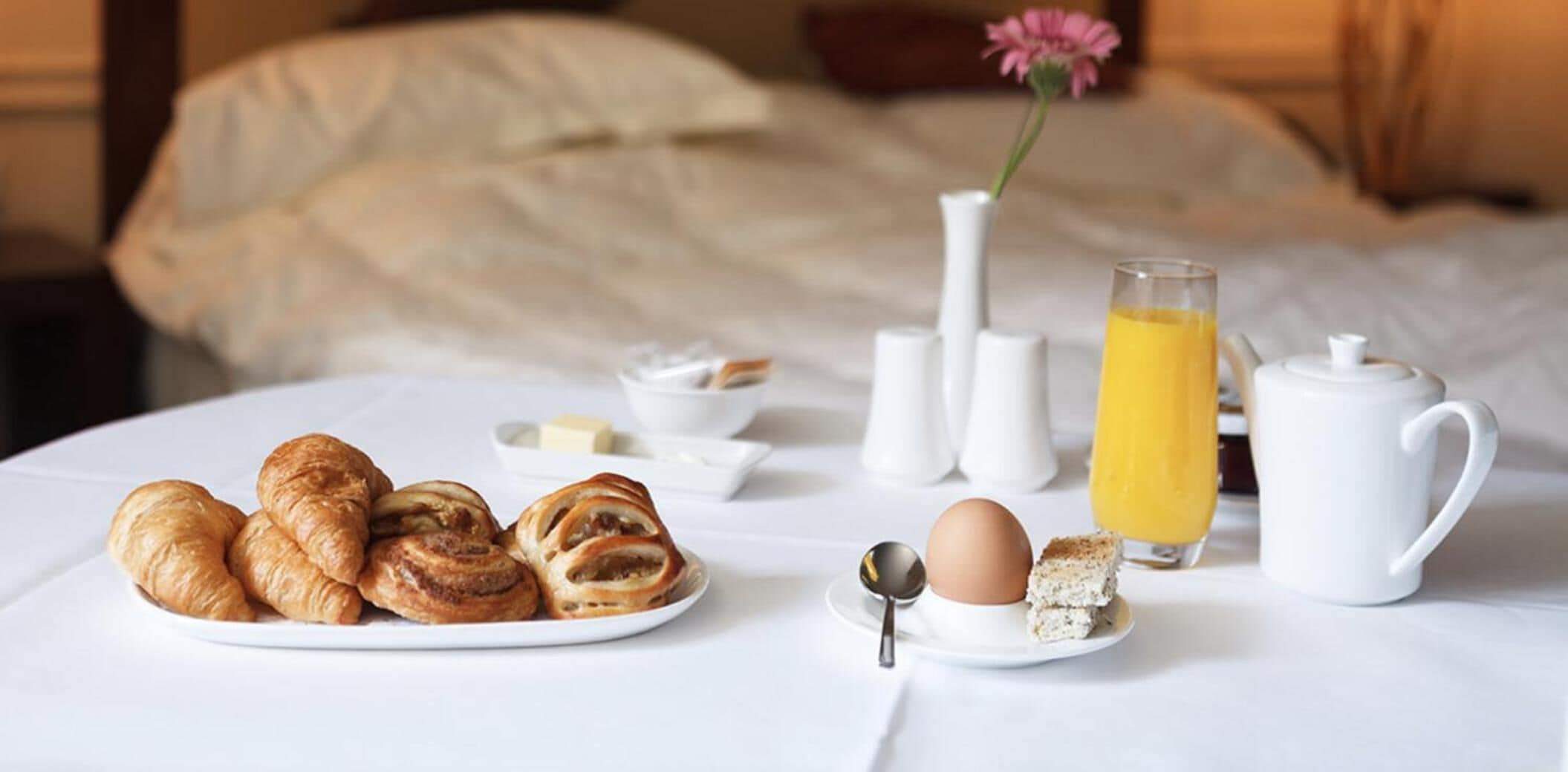 A breakfast setup on a bed with croissants, a glass of orange juice, a teapot, and a plate with a boiled egg and toast.