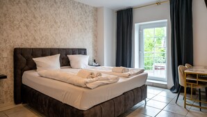 A bedroom with a large bed, a bedside table, a chair, a window with curtains, and a view of greenery outside. - Hotel Vintage (Pulheim)