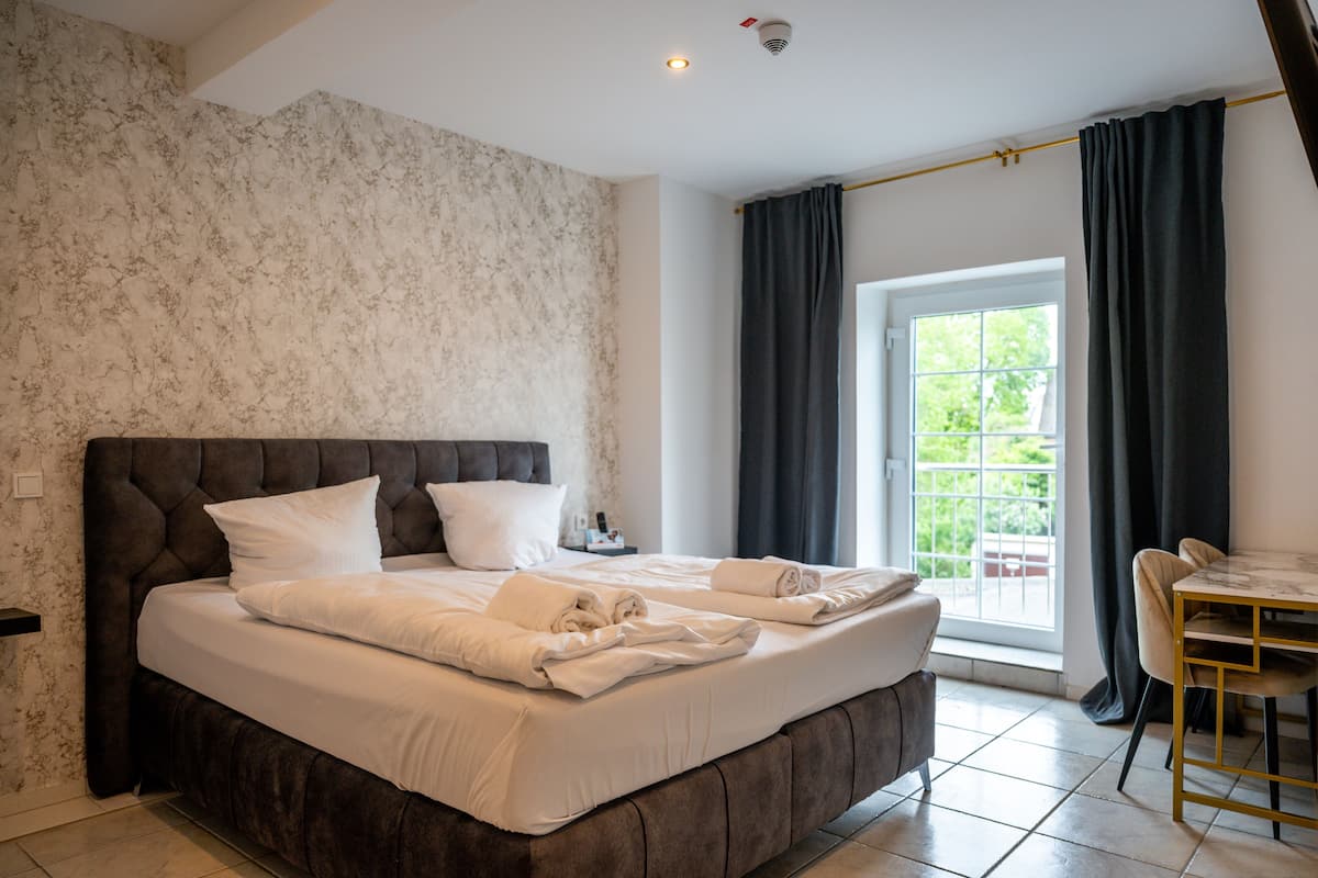 A bedroom with a large bed, a bedside table, a chair, a window with curtains, and a view of greenery outside.