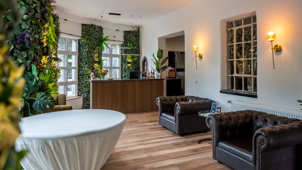 A modern interior with a wooden floor, two leather sofas, a round table, and a reception desk with a green wall and plants. - Hotel Vintage (Pulheim)