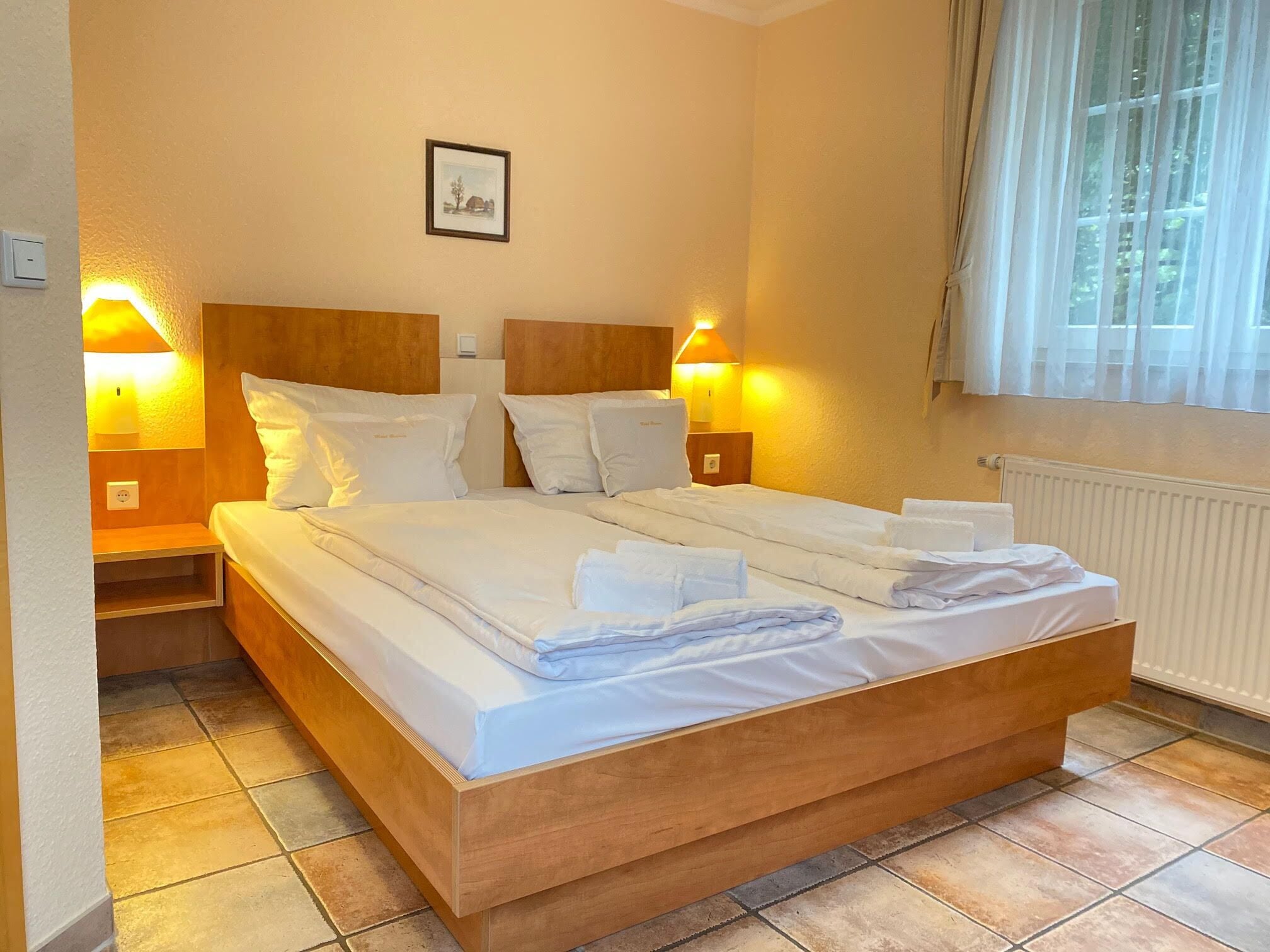 A double bed with white linens, two bedside tables with lamps, a framed picture on the wall, and a window with sheer curtains.