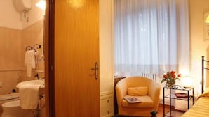 A bedroom with a bed, a chair, a bedside table, a window with curtains, and a bathroom with a toilet and sink. - Hotel PRime già Hotel Piccolo Ritz (Pistoia)