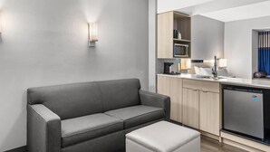 Een moderne hotelkamer met een grijze bank, een eettafel, en een kitchenette met een magnetron en een koelkast.