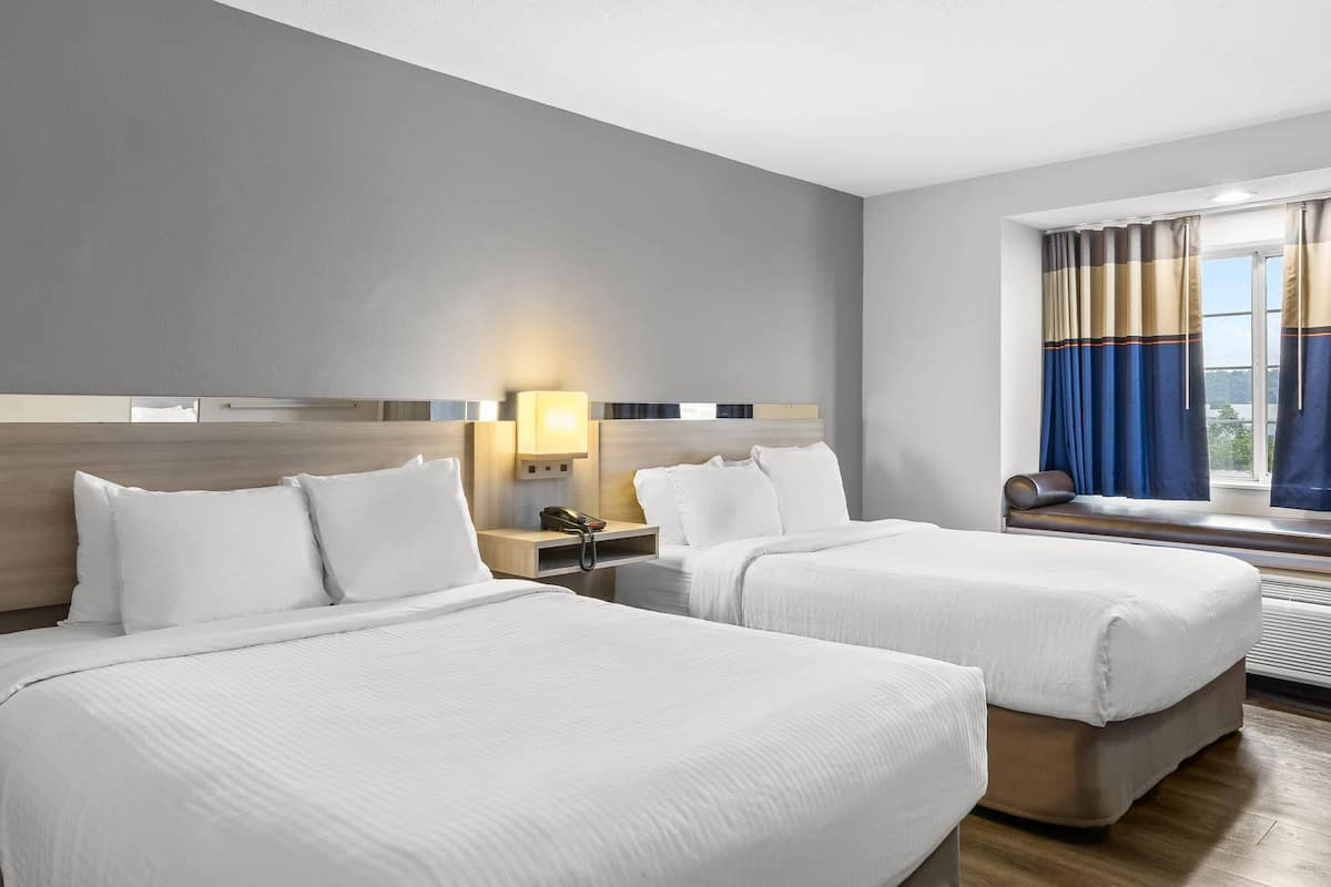 Habitación de hotel con dos camas, una mesita de noche con lámpara, una ventana con cortinas y vistas al exterior.